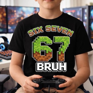 May include: Black t-shirt with the text "SIX SEVEN 67 BRUH" in a pixelated design. The numbers are green and brown, resembling a video game aesthetic. The person is holding a black video game controller.