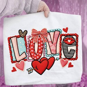 May include: White fabric with the word "LOVE" embroidered in a colorful, Valentine's Day theme. The letters are decorated with hearts, bows, and patterns. A pair of red hearts pierced by an arrow is also featured.