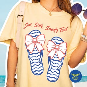 Puede incluir: Una camiseta amarilla clara con el texto "Sun, Salt, Sandy Toes" en escritura roja. La camiseta presenta un gráfico de dos chanclas azules y blancas, cada una adornada con un gran lazo rojo y blanco. El diseño es un gráfico con temática de playa.