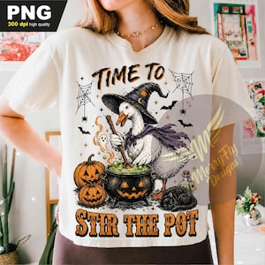 May include: A cream-colored t-shirt featuring a graphic of a white duck wearing a black witch hat and purple cape, stirring a black cauldron. The design includes two orange jack-o'-lanterns, a black cat, spiderwebs, and the text "TIME TO STIR THE POT."