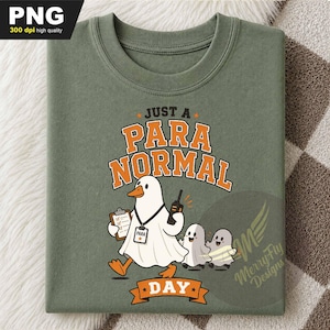 May include: An olive green t-shirt featuring a graphic of a duck wearing a white ghost costume, holding a clipboard and a walkie-talkie, with two smaller ghosts nearby. The text reads JUST A PARA NORMAL DAY with PARA on the duck's ID badge.