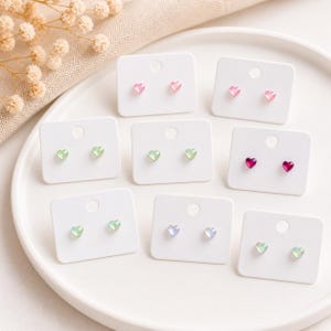 May include: A selection of heart-shaped stud earrings in shades of pink, green, and iridescent, presented on white cards. The earrings are arranged on a white plate, highlighting their delicate design and various colours.