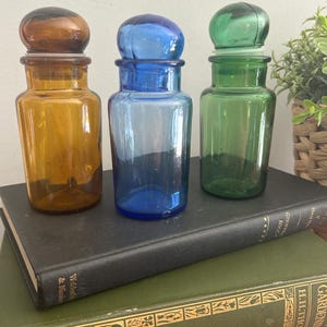 Peut inclure: Trois flacons d'apothicaire avec des bouchons en verre arrondis, chacun présentant une couleur translucide distincte : un ambre, un bleu et un vert. Ces récipients cylindriques en verre ont un col large et une finition lisse et polie.