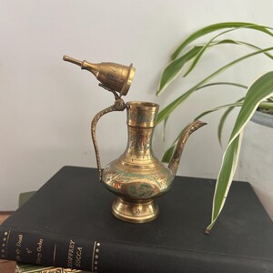 Peut inclure: Une cruche en laiton vintage avec une poignée et un bec décoratifs, ornée de motifs floraux et de feuilles gravés avec des accents verts et rouges. La cruche est surmontée d'un couvercle amovible. Elle est posée sur un livre noir.