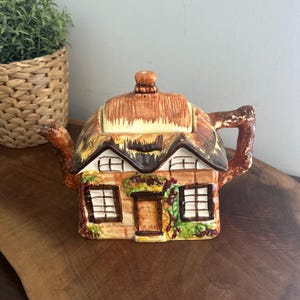 Peut inclure: Une théière en céramique en forme de cottage avec un toit de chaume, une porte centrale et deux fenêtres. L'extérieur présente des détails peints incluant des murs en briques brunes, des vignes vertes et une anse et un bec verseur texturés.