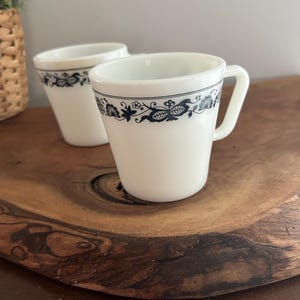 May include: Two white ceramic mugs with a dark blue floral pattern around the rim. The mugs have a handle and are placed on a wooden surface. The mugs are vintage and have a classic design.