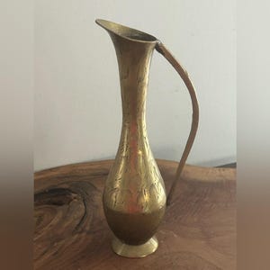 Peut inclure: Une cruche en laiton vintage avec un corps élancé et allongé et une poignée incurvée. La cruche présente des motifs floraux gravés et un bec évasé. La base est un petit piédestal circulaire. Le laiton a une teinte dorée chaude.