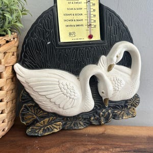 May include: A vintage bath thermometer featuring two white swans and a black frame. The thermometer, labelled "BATHOMETER", has temperature indicators. The swans rest on a base of gold and black leaves. The thermometer is likely to measure water temperature.
