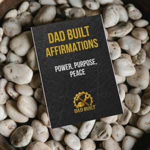Peut inclure: Un livre noir intitulé "DAD BUILT AFFIRMATIONS" avec des lettres dorées et un logo doré d'un marteau et d'une clé. Le livre est posé sur un lit de pierres blanches lisses.