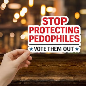 May include: A white sticker with red text that reads "STOP PROTECTING PEDOPHILES" above "VOTE THEM OUT" in blue text. The sticker is held in front of a blurred background.
