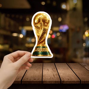 May include: A gold-colored FIFA World Cup trophy sticker with a green base. The sticker is held in a hand against a blurred background. The text "FIFA WORLD CUP" is visible on the base of the trophy. The sticker is on a wooden surface.
