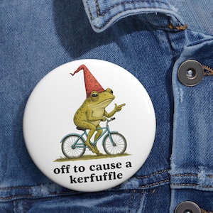 May include: A white button featuring a whimsical illustration of a frog riding a bicycle and wearing a red hat. The frog is pointing with its right hand. The text "off to cause a kerfuffle" is below the illustration.