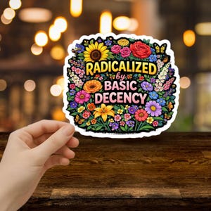 May include: A vibrant sticker featuring the words "RADICALIZED by BASIC DECENCY" within a floral design of sunflowers, roses, and other blooms. The sticker is held by a hand.
