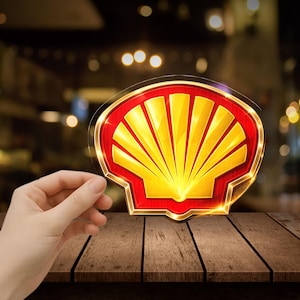 May include: A clear sticker featuring the Shell logo, a yellow scallop shell design with red trim. The logo is held in front of a blurred background. The sticker is on a wooden surface.