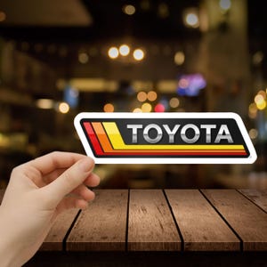 May include: A rectangular sticker with the word "TOYOTA" in silver lettering. The sticker has a black background with red, orange, and yellow stripes. The sticker is being held up by a hand.