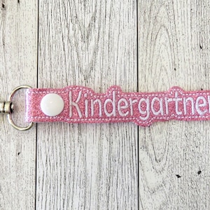 May include: Pink glitter keychain with a white button and the word "Kindergarten" embroidered in white.