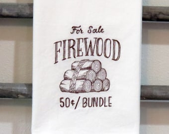 May include: White kitchen towel with brown embroidery that reads "For Sale Firewood 50¢/Bundle". The embroidery also includes a drawing of a stack of firewood.
