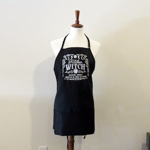 Op de afbeelding: Zwarte keukenschort met een wit ontwerp met de tekst "Kitchen Witch" met een ketel en maanfasen. De tekst eronder luidt "Magical things crafted in this kitchen."