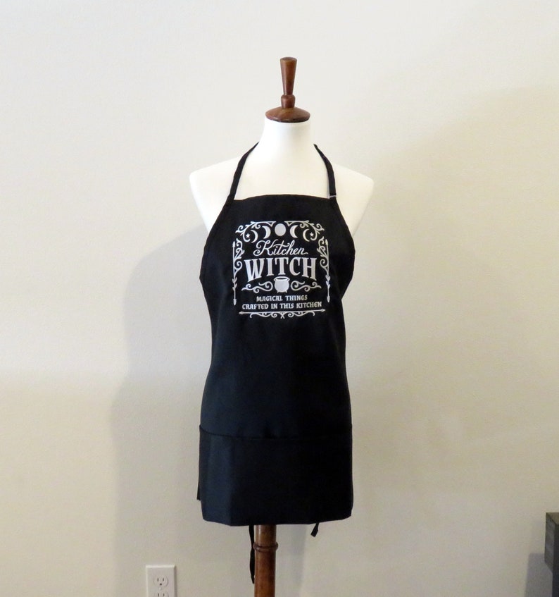 Op de afbeelding: Zwarte keukenschort met een wit ontwerp met de tekst "Kitchen Witch" met een ketel en maanfasen. De tekst eronder luidt "Magical things crafted in this kitchen."