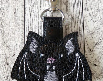 May include: Black bat-shaped keychain with white embroidered details. The bat has a pink nose and white fangs. The keychain includes a silver-colored clasp and a black leather strap with a black button.