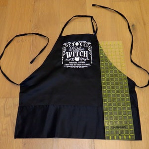 Op de afbeelding: Zwarte keukenschort met witte tekst die "Kitchen Witch Magical Things Crafted in This Kitchen" luidt.