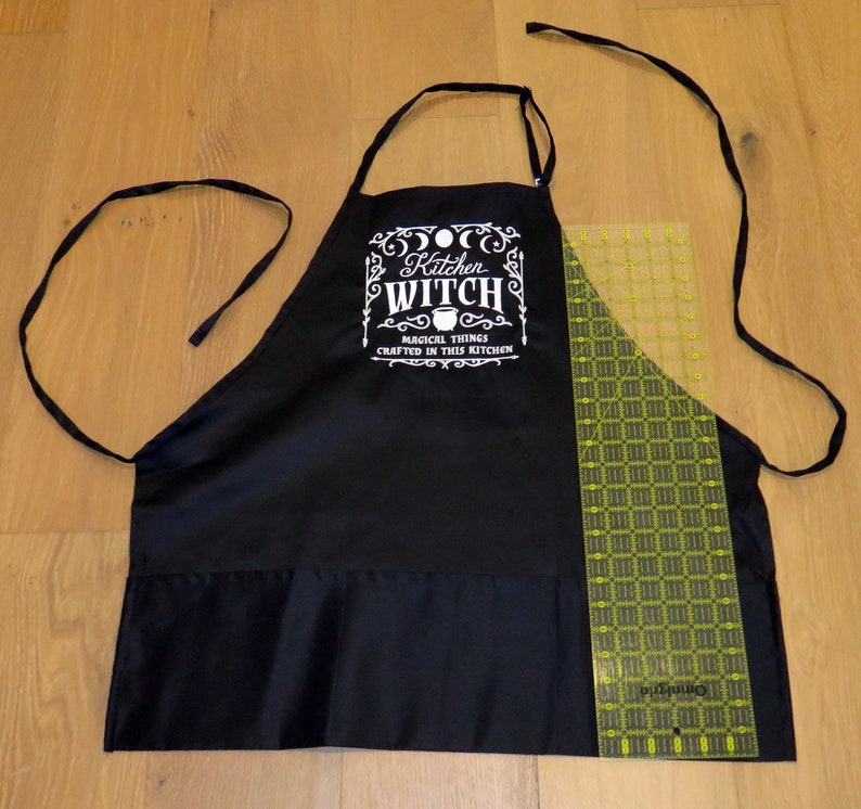 Op de afbeelding: Zwarte keukenschort met witte tekst die "Kitchen Witch Magical Things Crafted in This Kitchen" luidt.