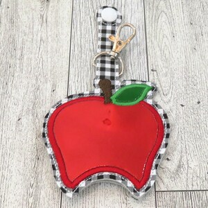 May include: A red apple-shaped keychain with a black and white checkered fabric backing. The keychain has a silver clip and a white button.