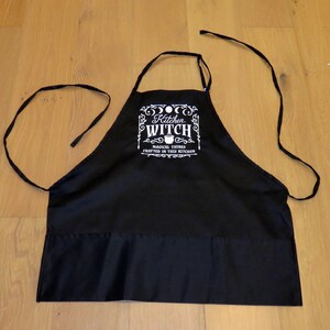 Op de afbeelding: Zwarte keukenschort met witte tekst die "Kitchen Witch Magical Things Crafted In This Kitchen" luidt. De schort heeft twee lange banden en een zak.