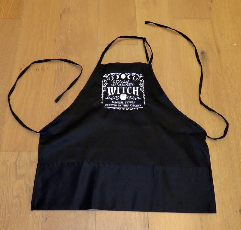Op de afbeelding: Zwarte keukenschort met witte tekst die "Kitchen Witch Magical Things Crafted In This Kitchen" luidt. De schort heeft twee lange banden en een zak.