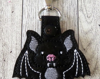 May include: Black bat-shaped keychain with white embroidered details on the wings and belly. The bat has a pink nose and small white fangs. A silver-colored clasp and a black leather strap are attached to the bat.