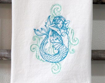 May include: A white kitchen towel with a blue and green embroidered mermaid design. The mermaid is depicted with flowing hair and a tail with scales. The design is surrounded by stylized waves.