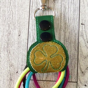 May include: A green fabric key chain with a gold glitter shamrock design. The key chain has two black buttons and holds multiple colorful hair ties.