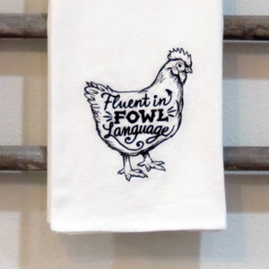 Puede incluir: Toalla de cocina blanca con una imagen bordada en negro de una gallina y el texto "Fluent in Fowl Language".