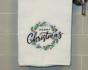 Puede incluir: Toalla de mano blanca con un diseño de corona de acebo verde y las palabras "MERRY Christmas" bordadas en escritura negra. La corona incluye bayas rojas y detalles azules. La toalla está colgada de una barra cromada.