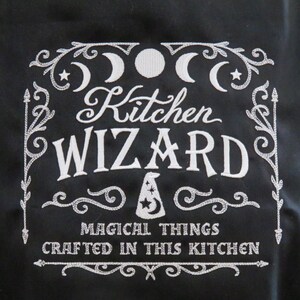 Puede incluir: Delantal negro con texto bordado blanco y elementos decorativos. El texto dice "Kitchen Wizard" con la frase "Magical Things Crafted in This Kitchen" debajo. El diseño incluye lunas, estrellas y un sombrero de mago.