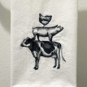 May include: A white kitchen towel with black embroidery of a cow, pig, and chicken stacked on top of each other.
