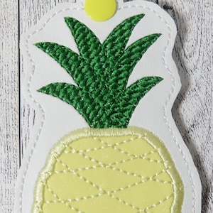 May include: A white leather keychain with a yellow and green embroidered pineapple design. The keychain has a silver metal clasp.