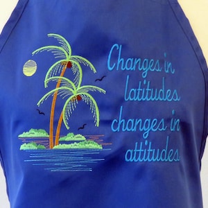 Pode incluir: Avental de cozinha azul com um desenho bordado de duas palmeiras e um pôr do sol. O texto "Changes in latitudes changes in attitudes" é bordado em azul no avental.