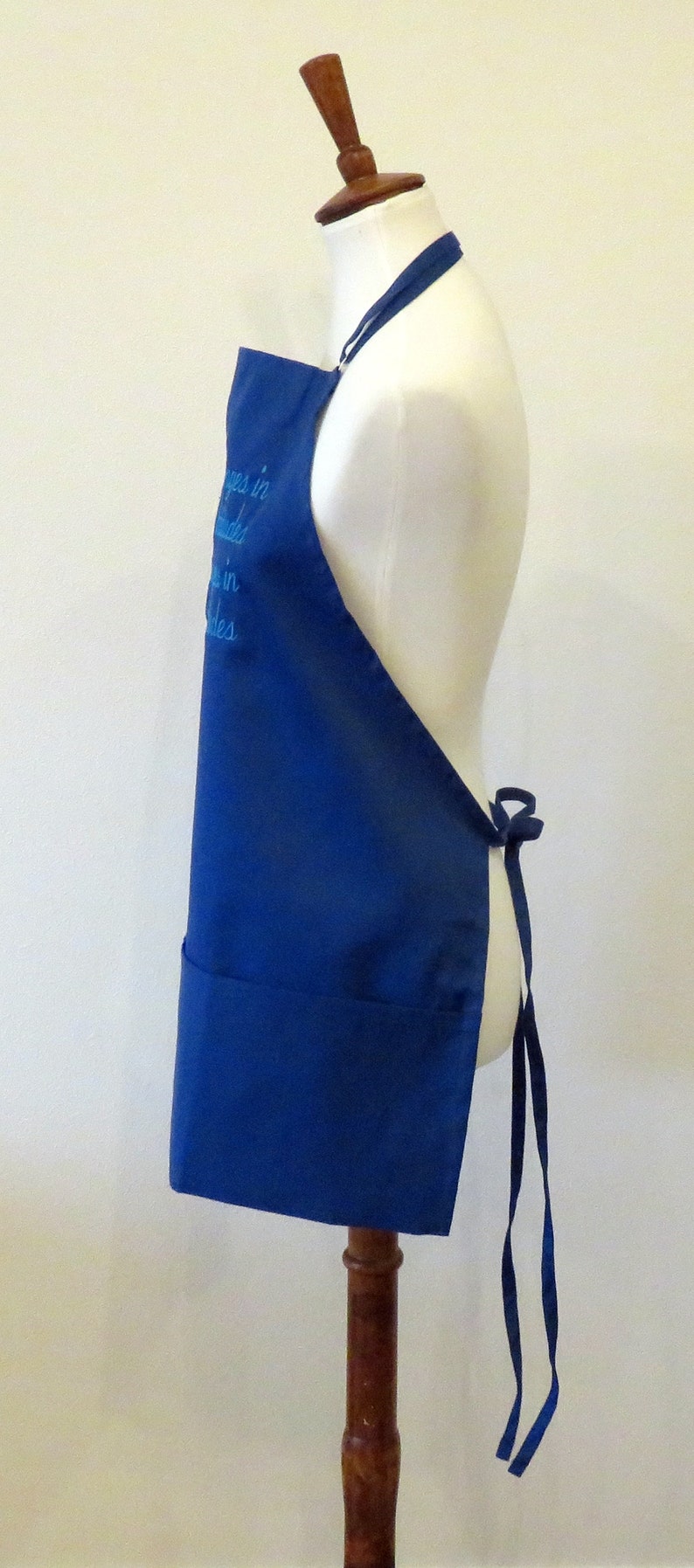 May include: A blue apron with a white embroidered saying "Happiness is homemade" on the front. The apron has a large pocket and ties at the waist.