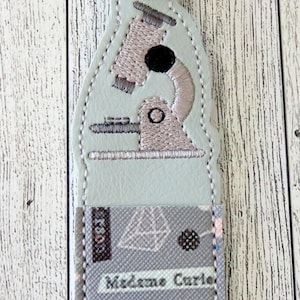 May include: A light blue keychain with a silver clasp. The keychain features an embroidered microscope design and the text "Madame Curie" on a gray patterned background. The keychain is a science-themed accessory.