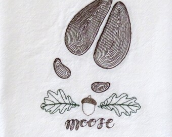 May include: Embroidered white kitchen towel featuring a brown moose footprint, two green oak leaves, and a brown acorn. The word "moose" is embroidered in brown cursive lettering below the design.