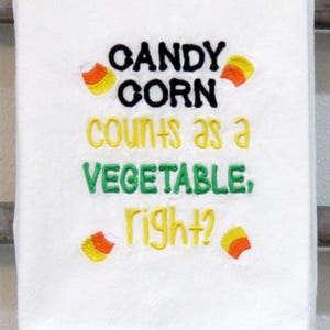 Peut inclure: Serviette blanche brodée avec le texte "CANDY CORN counts as a VEGETABLE, right ?" en fil noir, jaune et vert. Des graphiques de bonbons de maïs sont brodés autour du texte.