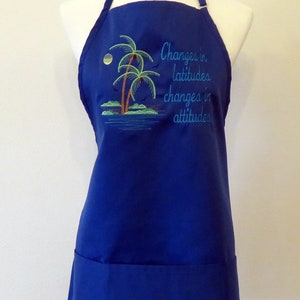 May include: A blue apron with a pocket and adjustable straps. It features an embroidered design of palm trees and the text "Changes in latitudes, changes in attitudes."