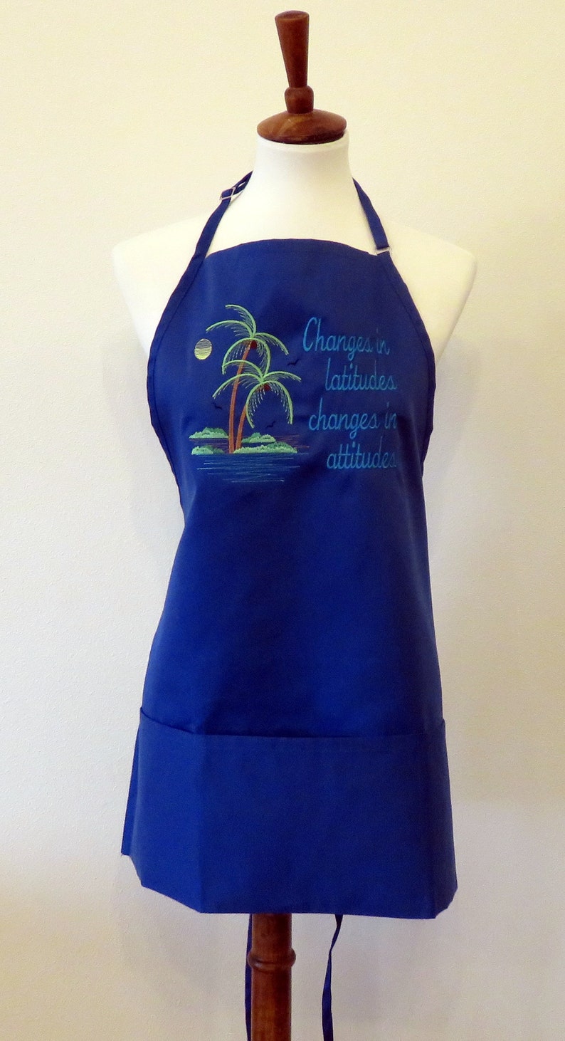 May include: A blue apron with a pocket and adjustable straps. It features an embroidered design of palm trees and the text "Changes in latitudes, changes in attitudes."