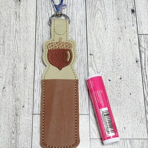 May include: A brown and tan lip balm holder with an acorn design, attached to a silver keychain. A pink lip balm tube is next to the holder. The holder is made of faux leather and has a stitched border.