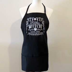 Op de afbeelding: Zwarte keukenschort met een wit ontwerp met de tekst "Kitchen Witch" met een ketel en de tekst "Magical things crafted in this kitchen".