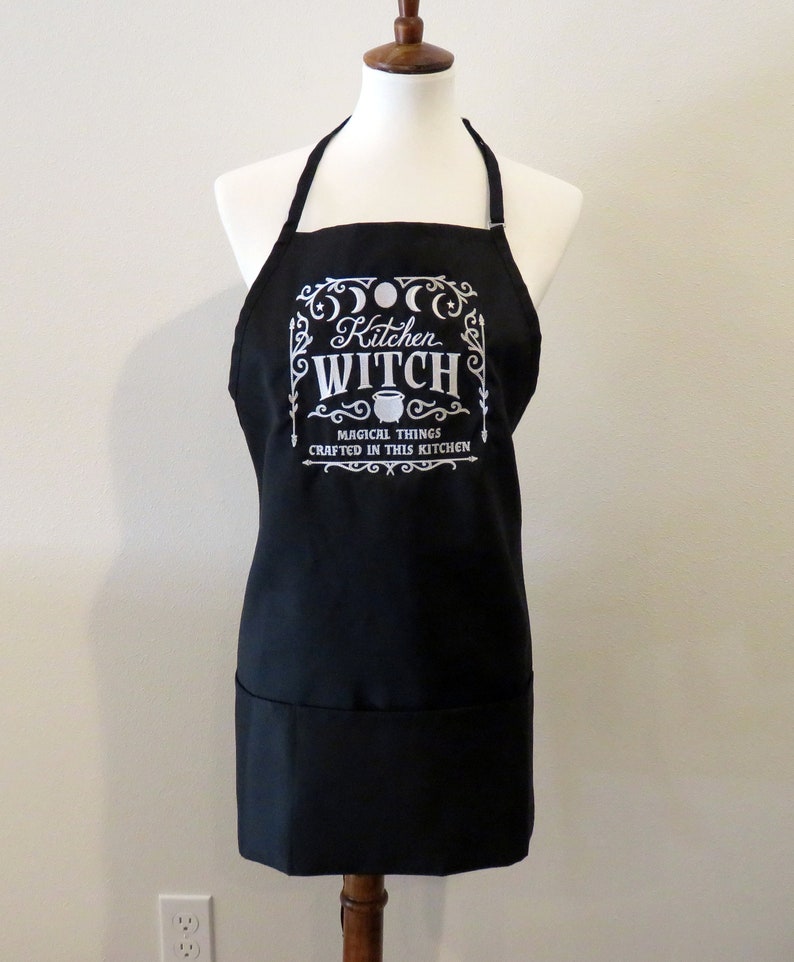 Op de afbeelding: Zwarte keukenschort met een wit ontwerp met de tekst "Kitchen Witch" met een ketel en de tekst "Magical things crafted in this kitchen".