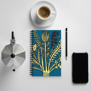 May include: A blue spiral bound notebook with a yellow and black botanical illustration of a plant with leaves and flowers. The notebook is on a white surface with a coffee cup, a pencil, and a smartphone.