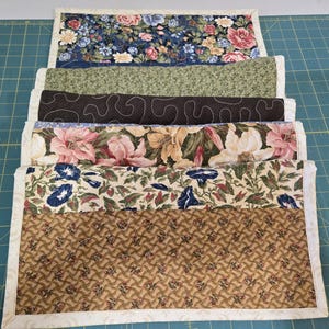 May include: A stack of six rectangular quilted placemats with floral and patterned designs. The placemats feature various colors, including blue, green, brown, pink, and beige. Each placemat has a white border. The top placemat has a blue floral pattern.