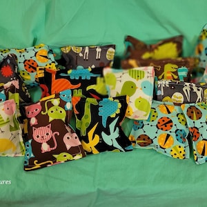 May include: A collection of 14 colorful fabric catnip toys. The toys are shaped like pillows and feature various animal and insect designs, including ladybugs, birds, cats, and dinosaurs. The toys are arranged on a green fabric background.